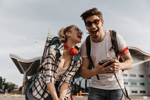 Blonde girl in plaid shirt and guy in white tee-shirt laugh near airport. Man holds retro camera. T