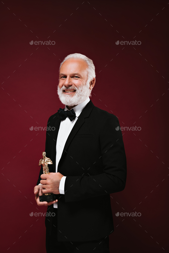 Cool man in classic style suit smiling and holding Oscar statuette. Stylish adult guy in white shir