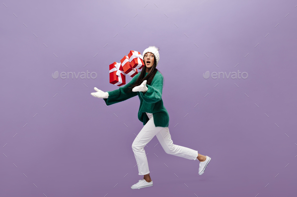 Tanned girl in white pants, stylish green woolen sweater, knitted hat, gloves throws up red gift bo