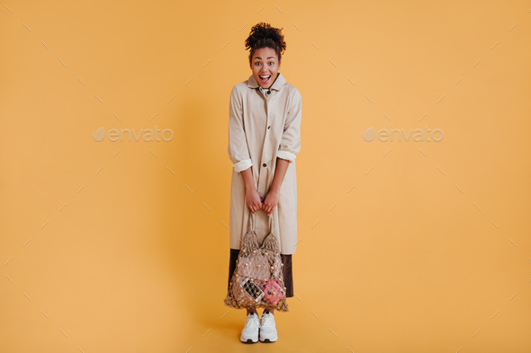 Shy african american girl holding string bag. Studio shot of pretty black woman in stylish trench c