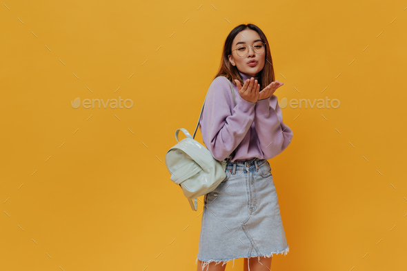 Pretty Asian woman in eyeglasses, denim skirt and purple sweater blows kiss and poses with mint bac