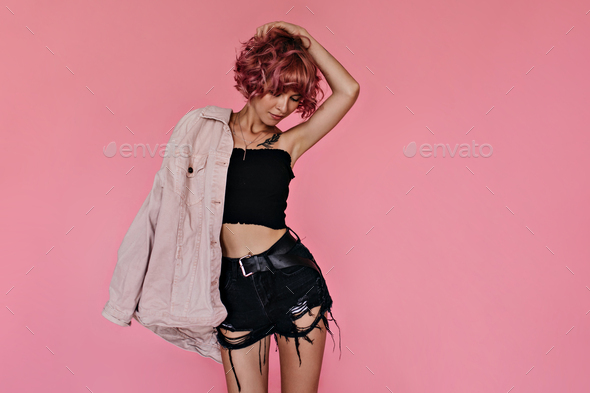 Curly young woman touches her pink hair and poses on isolated. Portrait of tanned girl in cropped t