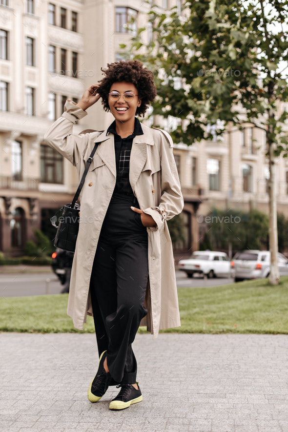 Joyful happy brunette curly dark-skinned woman in black pants, blouse and beige trench coat smiles