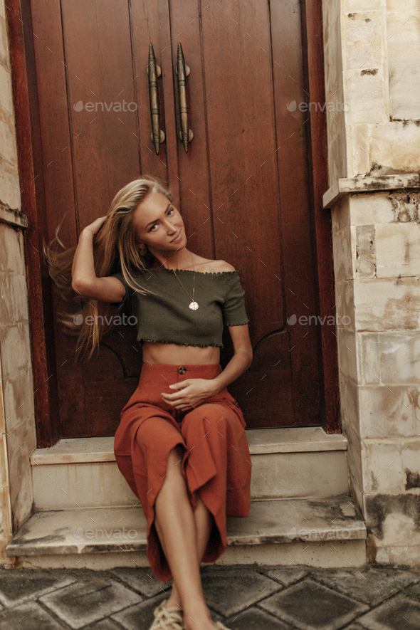 Tanned attractive blonde woman in khaki cropped t-short and red loose pants touches hair and sits n