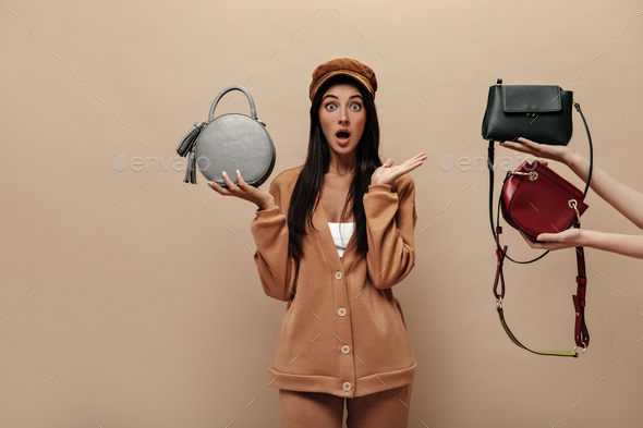 Surprised green-eyed woman holds round bag. Woman in cap and beige suit poses on isolated backgroun