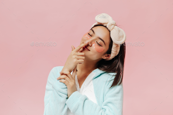 Brunette pretty woman in blue pajamas and soft beige headband touches nose. Cute Asian girl in slee