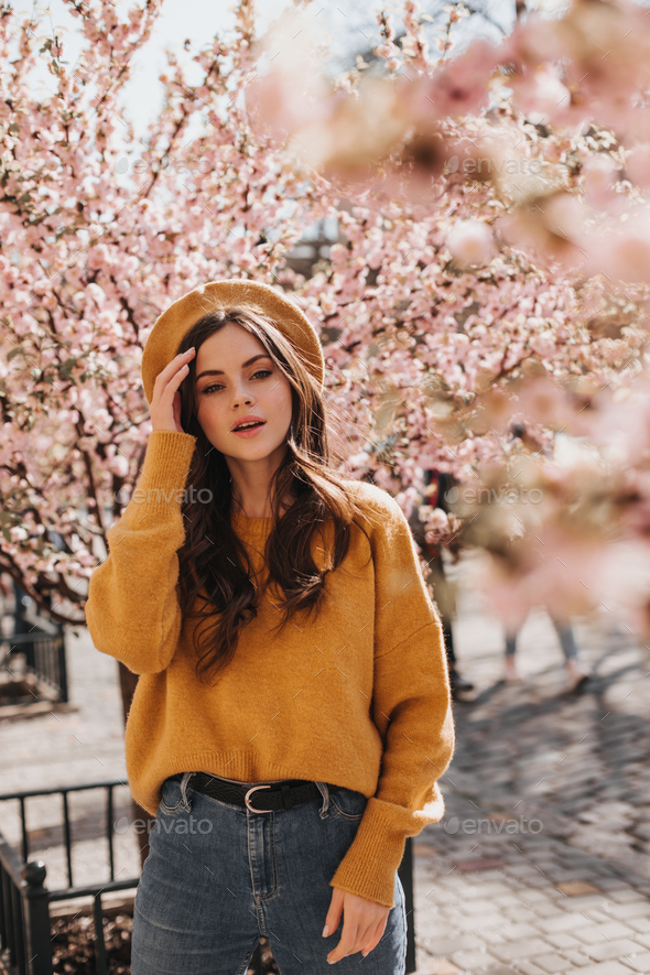 Brunette girl in stylish outfit and hat poses near sakura. Portrait of  woman in orange sweater, jea Stock Photo by look_studio