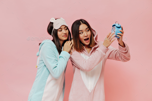 Asian girls in colorful cute pajamas look at blue alarm clock. Pretty women in sleep wear pose on p