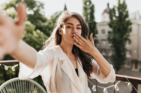 Curly woman in stylish shirt blowing kiss and taking selfie. Good-humored brunette lady in beige bl