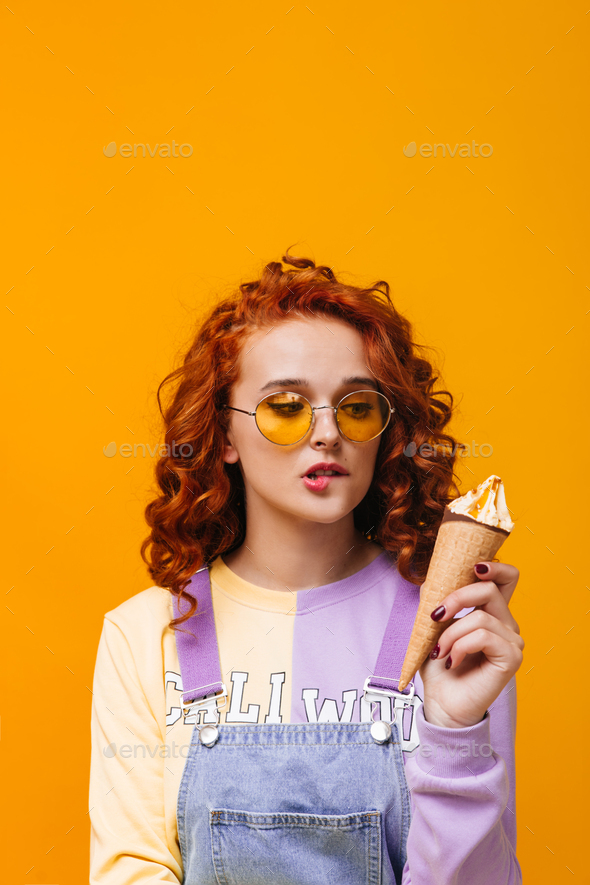 Attractive redhead girl in stylish sweatshirt and yellow glasses bites her lip and looks at ice cre