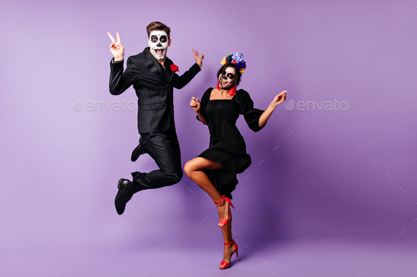 Full-length portrait of european couple dancing on purple background in zombie costumes. Funny youn