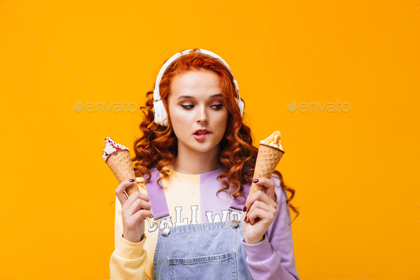Hungry girl with red hair bites her lip and wants to eat ice cream on orange background