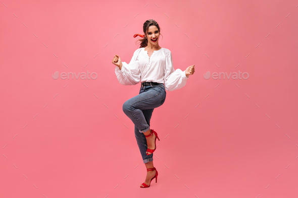 Joyful woman in jeans, white blouse dancing on pink background. Modern girl with red lipstick and i