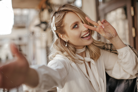 Cool beautiful blonde woman in white stylish blouse and pearl jewelry smiles, shows peace sign and