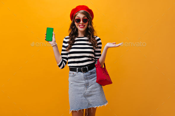 Pretty woman in sunglasses and red beret demonstrates smartphone. Attractive girl in denim skirt wi