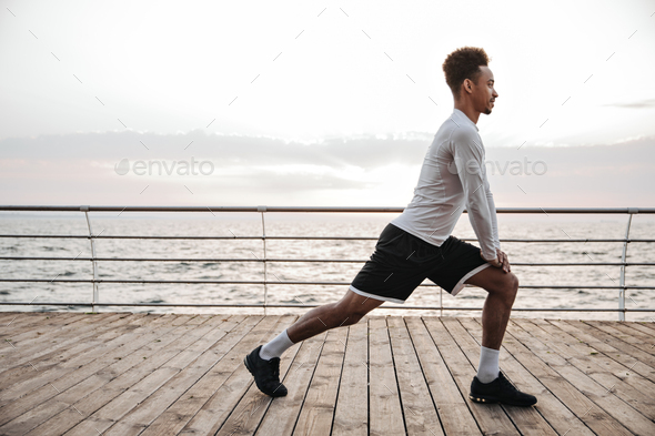 Strong active curly dark-skinned man in black shorts and white long-sleeved t-shirt squats, works o