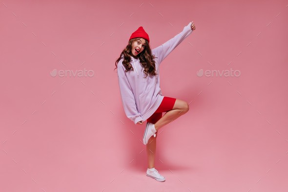 Brunette curly woman in red bright hat, purple hoodie and cycling shorts smiles widely and moves on