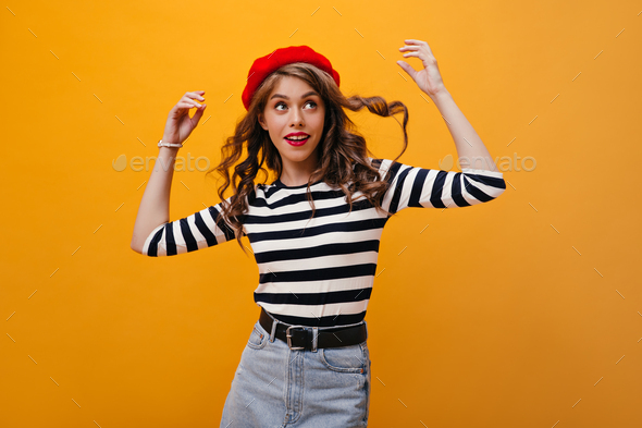 Pretty lady plays her curly hair on orange background. Fanny young woman in red beret, striped blou