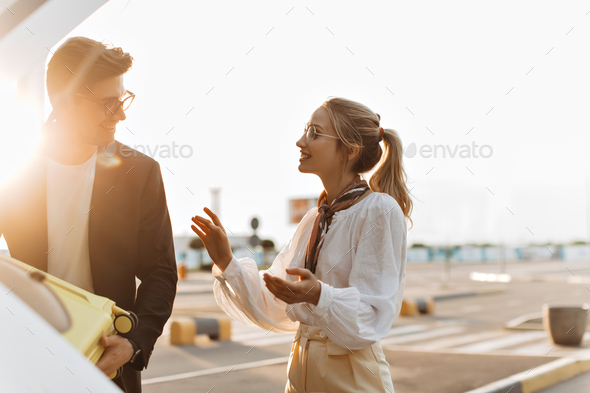 Happy blonde woman in white blouse talks with boyfriend outside. Charming man in black jacket and s