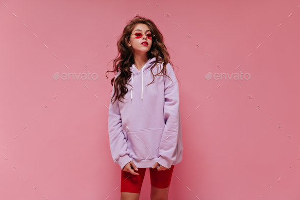 Charming woman in red cycling shorts and purple hoodie poses on pink background. Young girl in sung