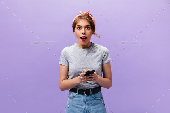 Shocked lady poses with black phone. Young fashionable woman in grey t-shirt and trendy skirt with