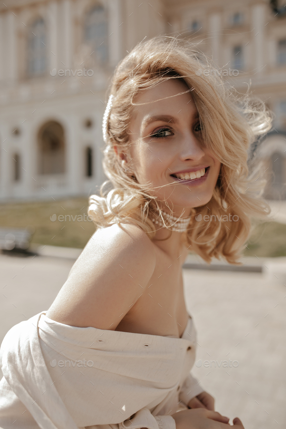 Cheerful blonde woman in stylish white blouse smiles widely outside. Happy curly lady in pearl neck
