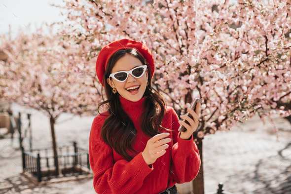 Woman in high spirits poses with phone near cherry blossoms. Portrait of lady in red beret, casheme