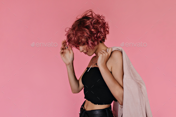 Short-haired curly woman in black cropped top and shorts holds denim light pink jacket. Cool tanned