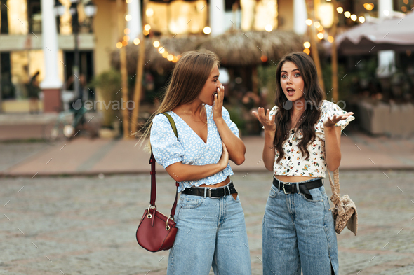 Curly brunette woman in white floral top and jeans talks to her friend and tells her secrets. Blond