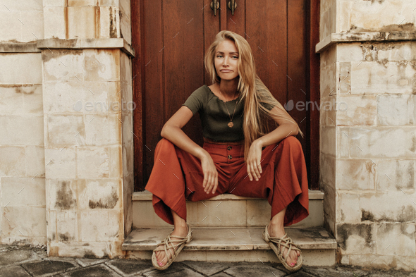 Cool young blonde sad long-haired girl in loose red pants and khaki t-shirt sits in funny pose near