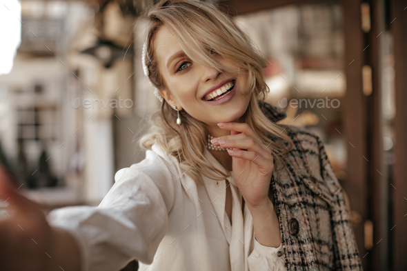Happy young blonde woman in white blouse and tweed checkered coat smiles sincerely and takes selfie