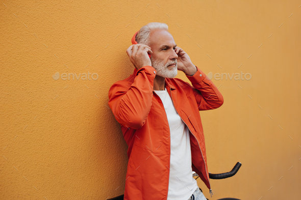 Adult man in headphones listening to music on orange background. Stylish gray-haired guy in white T