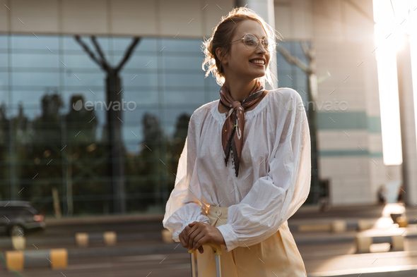 Pretty charming blonde woman in white blouse smiles sincerely. Happy girl in beige pants poses with