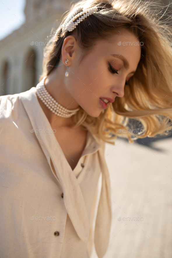 Charming curly blonde woman in pearl jewelry and beige blouse looks down, walk outside. Young girl