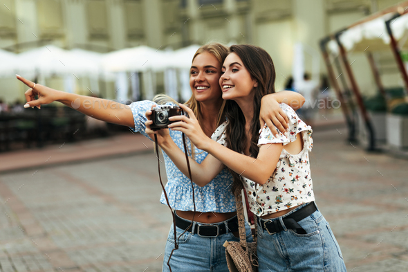 Cheerful brunette and blonde girlfriends in floral cropped blouses and denim pants smiles and walks