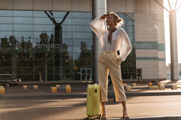 Stylish blonde woman in white blouse and beige pants poses with yellow suitcase outside. Portrait o