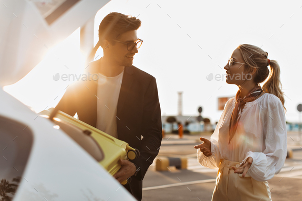 Brunette man in black jacket and blonde woman in white blouse smile and talk outside. Handsome guy