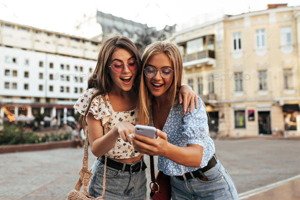 Happy young tanned girls in colorful sunglasses and stylish cropped blouses look at phone screen an