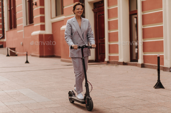 Fashionable lady in suit and eyeglasses rides electric scooter. Young  stylish woman dressed in grey Stock Photo by look_studio