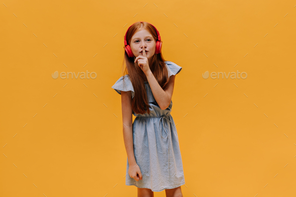 Redhead child asks to keep secret and to be silent. Little girl in blue dress listens to music on i
