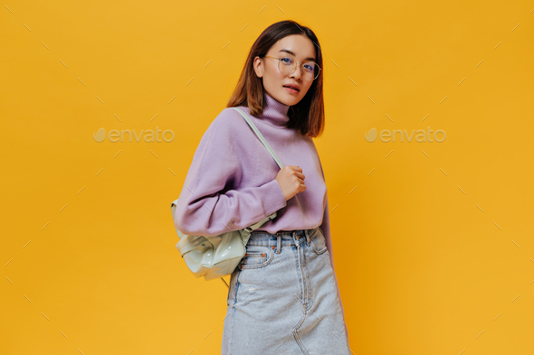 Portrait of tanned short-haired young woman in eyeglasses, denim skirt and purple sweater looks int