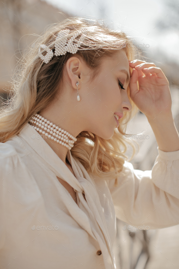 Pretty blonde curly woman in pearl necklace and earrings touches face. Happy girl in white blouse p