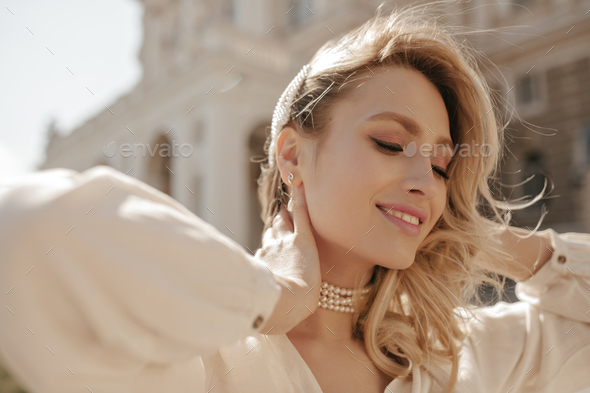 Cheerful blonde curly woman in pearl necklace and white elegant blouse smiles sincerely, looks down