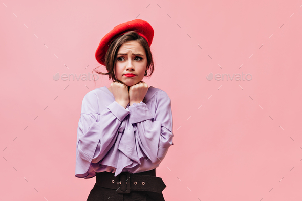 Lovely young girl makes innocent face. Portrait of lady in beret and stylish blouse on pink backgro