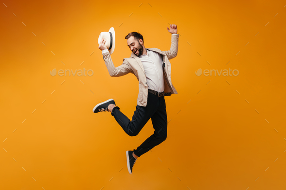 Happy man in suit takes off hat and jumps on orange background.