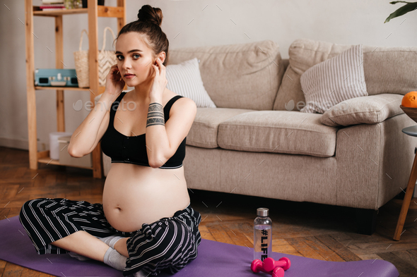 Blue-eyed brunette pregnant woman in black cropped top and striped pants looks into camera in livin