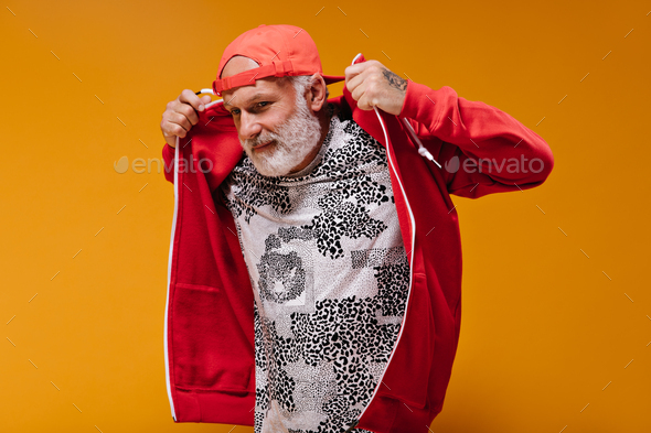 Stylish man in red cap puts on hoodie. Fashionable adult guy with grey hair in leopard print t-shir