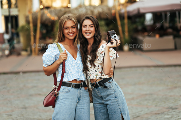 Tanned curly brunette woman in loose denim pants and floral blouse holds retro camera and poses wit