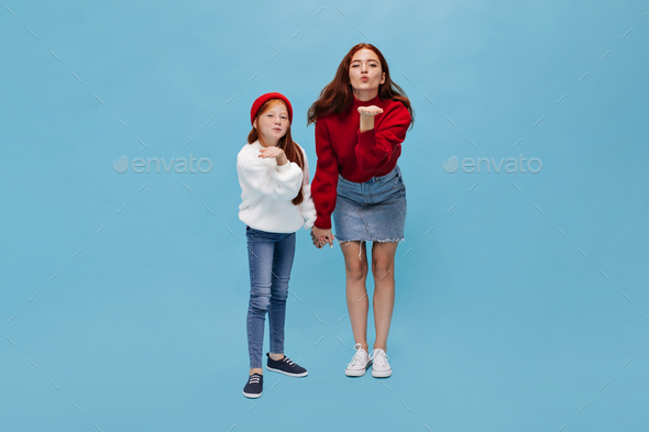 Stylish two sisters in stylish outfit blows kisses on blue background. Red haired girl in white swe