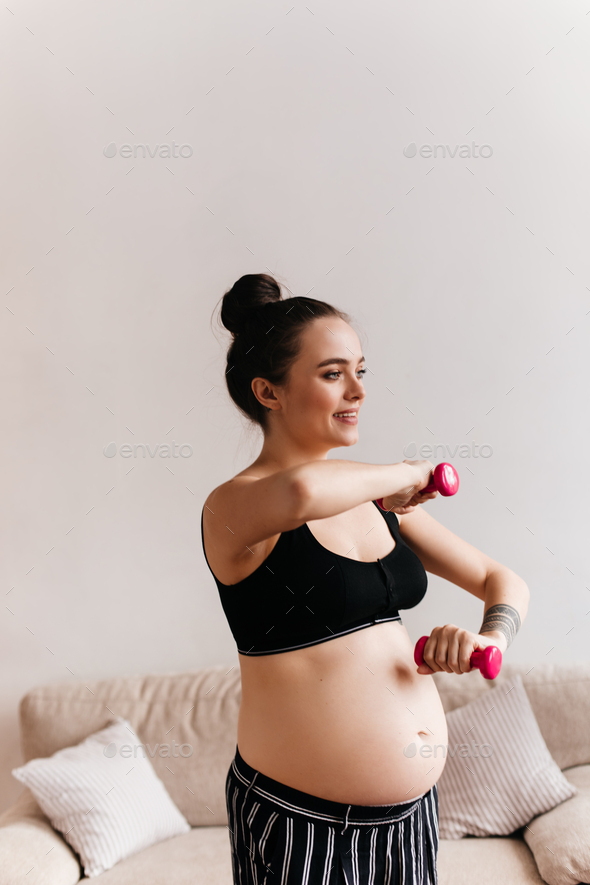 Smiling tanned woman in black cropped top and striped pants doing sport in living room. Pregnant la